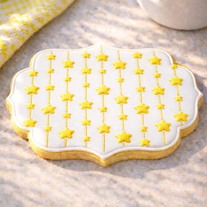 May include: A star-shaped cookie with a white icing background and yellow star decorations. The cookie has a decorative edge and is decorated with vertical lines of yellow stars.