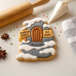 May include: A decorated gingerbread cookie shaped like a house with a brown door and the words "The Mean One's Go Away!" and "Unwelcome". The cookie is decorated with white icing to resemble snow and is on a white surface.
