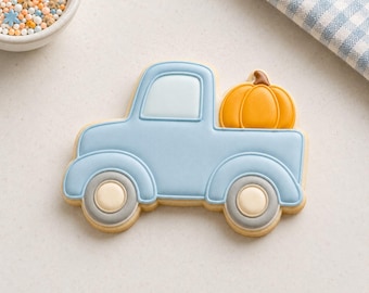 Può includere: Un biscotto a forma di camion decorato con glassa azzurra, con una zucca arancione brillante nel cassone. Le ruote sono dettagliate con glassa grigia e color crema, e il finestrino è riempito con una glassa di colore azzurro pallido.