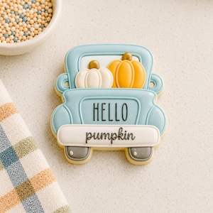 Puede incluir: Una galleta con forma de camión azul claro decorada con glaseado. La caja del camión lleva una calabaza blanca y una naranja. El texto HELLO está impreso en negro en la carrocería y la palabra pumpkin está escrita en negro en el parachoques.
