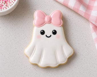May include: A ghost-shaped sugar cookie decorated with smooth white icing, featuring a light pink icing bow on top, two black circular eyes, a small black curved mouth, and soft pink blush accents on the cheeks, finished with a defined icing border.
