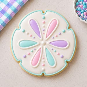May include: A round, sand dollar-shaped sugar cookie decorated with white royal icing and a central flower pattern featuring alternating pastel purple, pink, and teal teardrop shapes, accented with small white, pink, and purple icing dots.