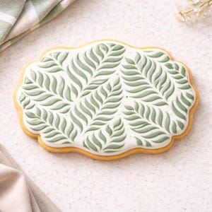 May include: A decorated cookie with a white base and a green leaf pattern. The cookie has a scalloped edge and a golden-brown border. The leaf design is a repeating pattern of stylized leaves.