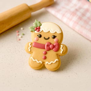 May include: A decorated gingerbread man cookie with a white icing head, a red bow tie, and a red scarf. The cookie has a smiling face, pink cheeks, and is decorated with holly leaves and berries. A rolling pin and a pink and white checkered cloth are in the background.