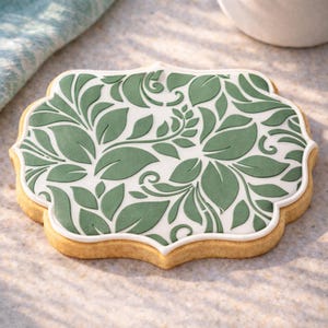 Puede incluir: Una galleta decorada con una base blanca y un patrón de hojas verdes. La galleta tiene un borde festoneado y un borde blanco. La galleta es de color marrón claro y parece recién horneada. El diseño recuerda un tema botánico.