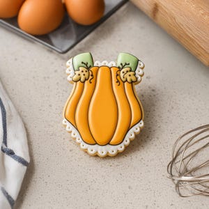 Pode incluir: Um biscoito em forma de abóbora decorado com glacê laranja, detalhes verdes e uma borda branca ondulada. O biscoito está em uma superfície clara, com utensílios de cozinha e ovos ao fundo. Uma guloseima festiva para o outono.