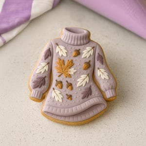 Puede incluir: Una galleta con forma de suéter lavanda decorada con detalles otoñales. La galleta presenta un cuello alto, mangas largas y está adornada con hojas de arce, bellotas y otros diseños de hojas en tonos blancos, marrones y morados.