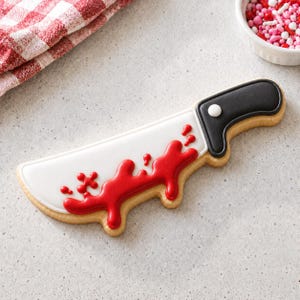 Puede incluir: Una galleta con forma de cuchillo decorada con glaseado blanco en la hoja y un mango negro. La hoja presenta un diseño de glaseado rojo brillante que se asemeja a salpicaduras de sangre. Un punto de glaseado blanco está en el mango negro.