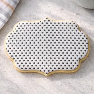 May include: A decorated sugar cookie with a white base and a pattern of evenly spaced black dots. The cookie has a decorative, scalloped edge and a light yellow border. The cookie is on a light-colored surface.