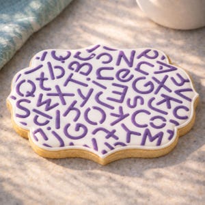 Peut inclure: Un biscuit sablé à bords festonnés recouvert d'un glaçage blanc lisse, orné d'un motif répétitif de lettres majuscules violettes. Le biscuit présente une croûte cuite brun doré clair visible sur tout le contour extérieur.