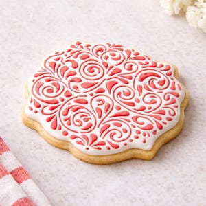 Puede incluir: Una galleta decorada con una base blanca y un diseño floral rojo en espiral. La galleta tiene un borde festoneado marrón claro y está sobre una superficie blanca. El diseño es intrincado y detallado.
