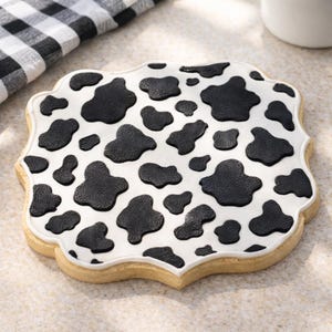 Puede incluir: Una galleta de azúcar decorada con una base blanca y manchas negras de vaca. La galleta tiene un borde festoneado y un borde blanco. La galleta está sobre una superficie de color claro.