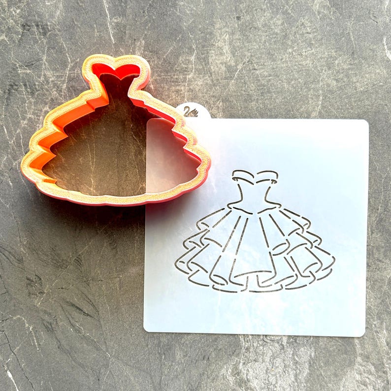 May include: A red and orange plastic dress-shaped cookie cutter next to a stencil with a dress design. The cookie cutter has a layered design, and the stencil features a detailed outline of a tiered dress.