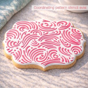 May include: A decorated cookie with a white base and a pink, swirling pattern. The cookie has a scalloped edge and the text "Coordinating pattern stencil avial" is visible in the background. The cookie is on a light-colored surface.