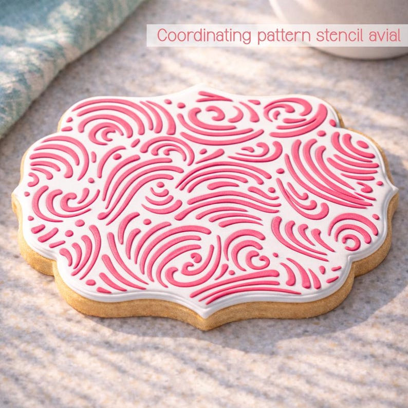 May include: A decorated cookie with a white base and a pink, swirling pattern. The cookie has a scalloped edge and the text "Coordinating pattern stencil avial" is visible in the background. The cookie is on a light-colored surface.