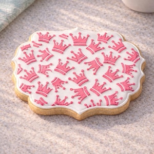 May include: A decorated biscuit with a white icing design featuring multiple pink crowns. The biscuit has a scalloped edge and a light brown colour. The crowns are evenly spaced across the white icing.