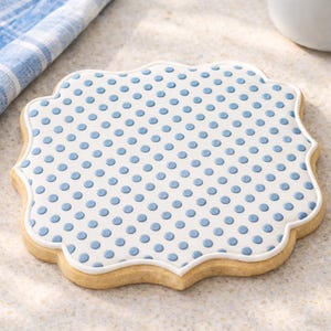 May include: A decorated sugar cookie with a white background and blue polka dots. The cookie has a scalloped edge and a white border. The cookie is on a light-colored surface.