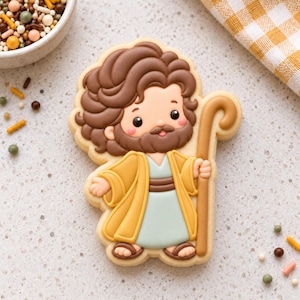 May include: A sugar cookie shaped like a person with brown hair and a beard, wearing a yellow robe over a light blue tunic. The figure holds a tall brown shepherd's staff in one hand and has pink cheeks, rendered in detailed icing.