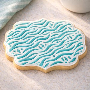 May include: A decorated sugar cookie with a white base and a turquoise wave pattern. The cookie has a scalloped edge and is decorated with small fish and dot details. The cookie is light brown.