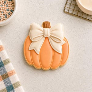 May include: A pumpkin-shaped sugar cookie decorated with smooth orange icing and a large, cream-colored icing bow centered at the top. The cookie features a small brown icing stem and defined vertical ridges, creating a clean, flat surface design.