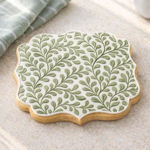 May include: A decorative cookie with a light brown base and a white, scalloped border. The cookie is topped with a detailed green leaf pattern. The design features a repeating pattern of leaves and vines, creating an elegant and natural aesthetic.