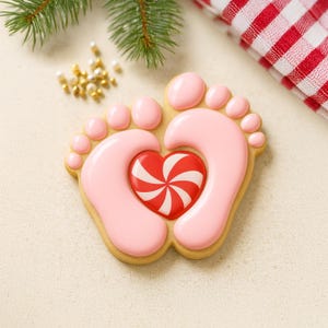 May include: A decorated sugar cookie in the shape of two baby footprints with a red and white heart in the center. The footprints are pink with a light brown outline. A sprig of evergreen and a red and white checkered cloth are in the background.