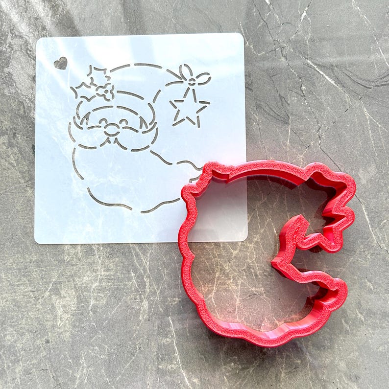 May include: A white stencil featuring a Santa Claus design with a heart, holly, and star cutouts. A red, scalloped-edge cookie cutter is next to the stencil. The items are on a gray surface. Ideal for holiday baking.
