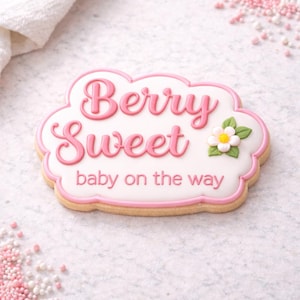 May include: A decorated sugar cookie in a cloud shape with pink frosting and the words "Berry Sweet baby on the way." The cookie has a white background, pink border, and a small flower.