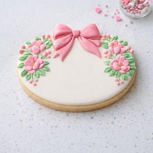 May include: A round sugar cookie decorated with a pink bow and floral design. The cookie features a white icing base with pink flowers, green leaves, and pink berries. The bow is centered at the top, and the flowers are arranged on either side.