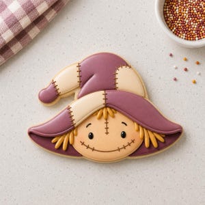 Puede incluir: Una galleta con forma de espantapájaros decorada con glaseado real. La galleta presenta un sombrero morado flexible con parches color crema, cabello amarillo y una cara sonriente con detalles de costuras en glaseado negro sobre fondo beige.