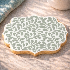 May include: A scalloped-edge cookie featuring a white base decorated with a repeating sage green teardrop or leaf-like pattern. The cookie has a light golden-brown baked crust visible along the edges, creating a distinct contrast with the icing.