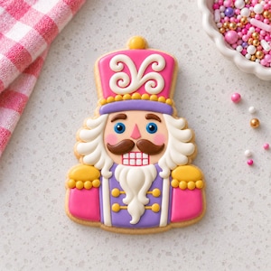 May include: A nutcracker-shaped sugar cookie decorated with royal icing in shades of pink, purple, white, brown, and yellow. The cookie features a pink hat with white swirls, blue eyes, a brown mustache, and a white beard and hair.