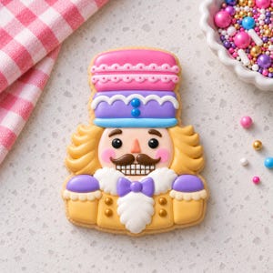 May include: A nutcracker-shaped sugar cookie decorated with royal icing. The cookie features a pink and purple hat, yellow hair, a brown mustache, a white beard, a purple bowtie, and a yellow jacket with gold buttons and purple accents.