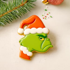 May include: A green cookie shaped like a character's face, wearing a red Santa hat with white trim. The cookie has a grumpy expression and is decorated with yellow, white, and orange icing. Christmas-themed food.