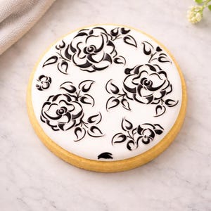 May include: A round cookie with a white icing top decorated with black floral designs. The cookie has a golden-brown edge and a detailed pattern of roses and leaves.