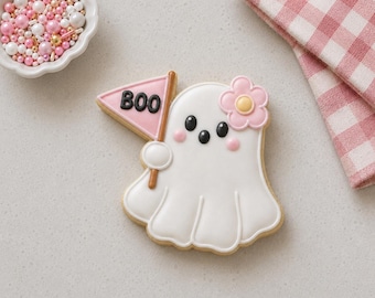 May include: A ghost-shaped sugar cookie decorated with white icing, black eyes, and pink cheeks. The ghost holds a pink triangular flag with the black text BOO and features a light pink flower with a yellow center on its head.