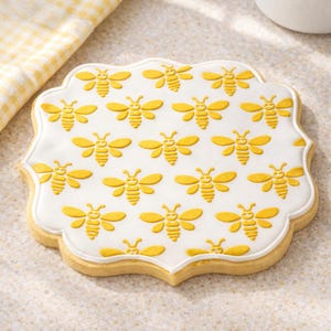 Puede incluir: Una galleta de azúcar decorada con una base de glaseado blanco. La galleta está adornada con un patrón de abejas amarillas. La galleta tiene un borde decorativo y una base marrón dorada. Un paño de cuadros amarillos y blancos está en el fondo.