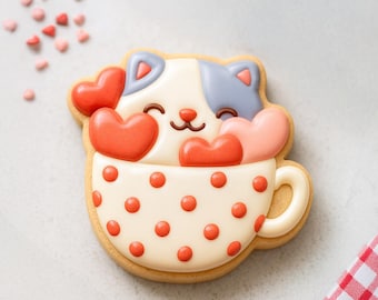 May include: A decorated sugar cookie in the shape of a cat in a teacup, with red heart-shaped candies. The cat has white and gray fur, with red hearts. The teacup is white with red polka dots.