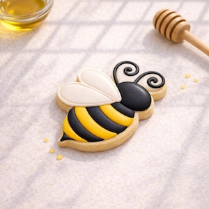 May include: A decorated bee-shaped cookie with black and yellow stripes, white wings, and black antennae. A small bowl of honey and a wooden honey dipper are in the background. The cookie is on a white surface.
