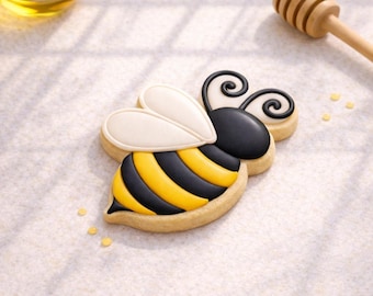 May include: A decorated bee-shaped cookie with black and yellow stripes, white wings, and black antennae. A small bowl of honey and a wooden honey dipper are in the background. The cookie is on a white surface.