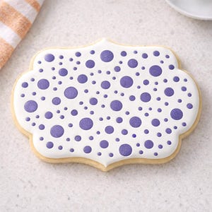 May include: A decorated sugar biscuit with a white base and purple polka dots. The biscuit has a scalloped edge and a light brown border. A striped orange and white tea towel is in the background.