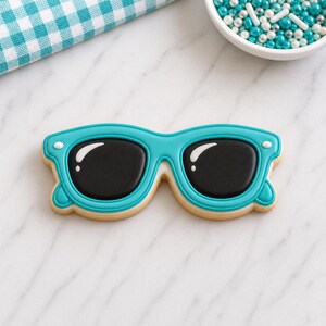 May include: A turquoise sunglasses-shaped cookie with black lenses and white accents. The cookie is decorated with a light tan icing border. A blue and white checkered napkin and a bowl of sprinkles are in the background.