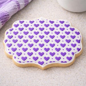 Puede incluir: Una galleta en forma de corazón con una cobertura blanca, decorada con un patrón de corazones y puntos morados. La galleta tiene un borde horneado de color marrón claro y un borde blanco. Un paño a cuadros morado y blanco está en el fondo.