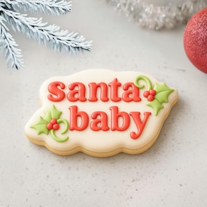 May include: A festive cookie with the words "santa baby" in red icing. The cookie is decorated with green holly leaves and red berries. The cookie has a light beige base and is a holiday treat.