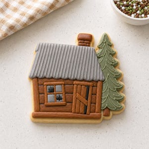 Könnte beinhalten: Ein hüttenförmiger Keks, verziert mit braunem Zuckerguss für die Blockwände und die Tür, einem grauen Wellblechdach und einem grünen Tannenbaum an der Seite. Der Keks hat ein Fenster mit grauen Scheiben und einen braunen Schornstein.