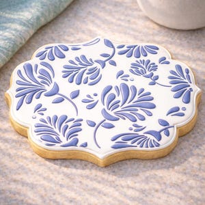 Puede incluir: Una galleta de azúcar decorada con una base blanca y un diseño floral azul. La galleta tiene un borde festoneado y un color marrón claro. El patrón floral está en relieve, dando un efecto 3D. La galleta es un dulce.