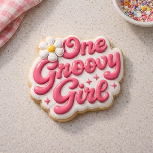 May include: A decorated sugar cookie with the words "One Groovy Girl" in pink frosting. The cookie has a white base with pink lettering and a daisy accent. Sprinkles and a pink and white checkered cloth are in the background.