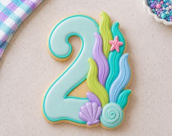 May include: A number two shaped sugar cookie decorated with light blue icing. The cookie features royal icing details including green and purple seaweed, a small pink starfish, a purple seashell, and a teal spiral seashell at the base.