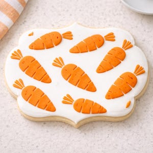 Puede incluir: Una galleta de azúcar decorada con una base de glaseado blanco. La galleta tiene un borde festoneado y está adornada con diseños de zanahorias naranjas. Las zanahorias tienen copas verdes y están dispuestas sobre la superficie de la galleta.
