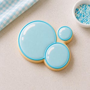 Puede incluir: Una galleta con forma de tres burbujas conectadas de distintos tamaños, decorada con glaseado azul claro liso y contornos en azul oscuro. Cada burbuja presenta un reflejo blanco de glaseado para un aspecto brillante y reflectante.